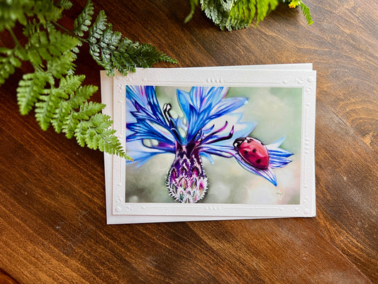 Focus: a card featuring soft pastel artwork of a ladybug on a blue flower. Background: a dark wooden desk and a fern. 